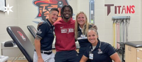 Group photo of three Beacon Athletic Trainers and IU South Bend athlete in training room with arms around each other's shoulders, smiling at camera; white female trainers wearing short-sleeved blue shirts, black male student wearing red sweatshirt with school logo.