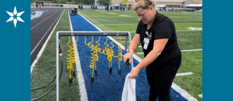 Female Beacon athletic trainer in black outfit, sprays off white towel with yellow hose, along sideline of football field.