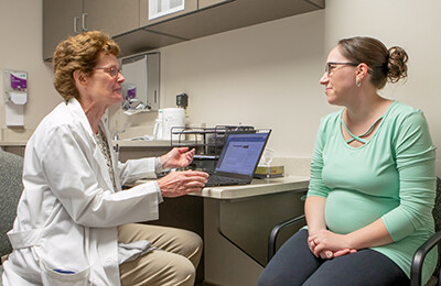 In medical exam room, seated female provider gestures with hands, facing seated pregnant patient.