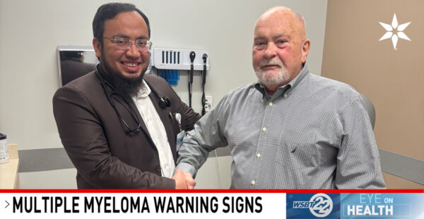 A doctor and a patient shake hands in an exam room, with medical equipment on the wall behind them and a banner below reading “Multiple Myeloma Warning Signs.”