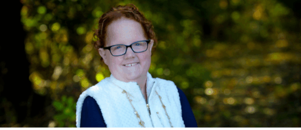 Photo of Genevieve Nikolai, a smiling, red-hairs young woman wearing a blue top and a white vest.