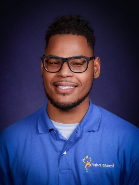 Professional portrait of Romie Granger wearing glasses and a blue Project SEARCH polo against a dark background.