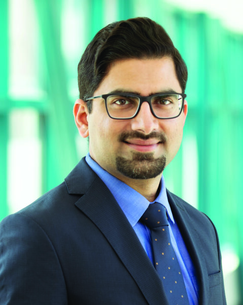 Muhammad Saeed, MD, a physician in a dark suit and glasses standing in a bright hospital corridor.