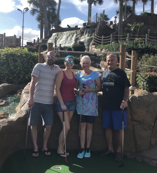 The Voorde family (Son Neal, Daughter Paige, Sarah and Fred) stand together on a mini-golf course, each holding a putter, posing in front of a rocky waterfall feature. 