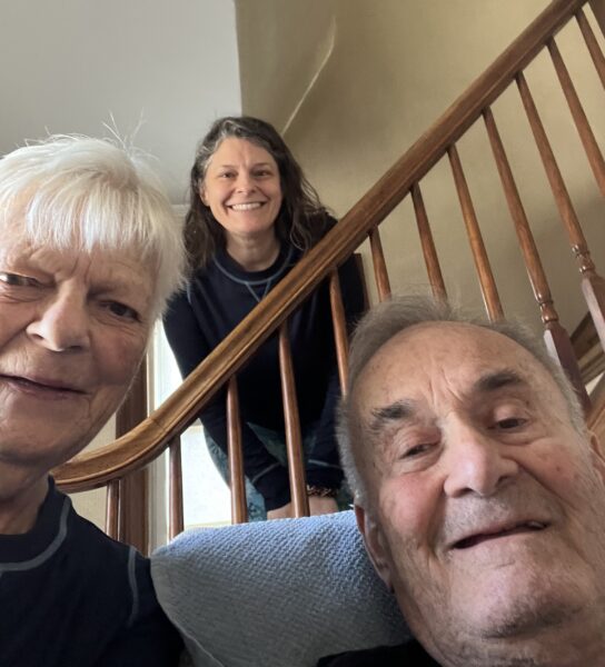 Sarah, Paige and Fred in 2025. Sarah and Fred sit together, with Paige leaning over the stair banister behind them. The family has broad and welcoming smiles.
