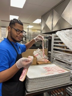 Romie Granger prepares bacon on trays in the Memorial Hospital kitchen, wearing gloves beside a rack, 2026.